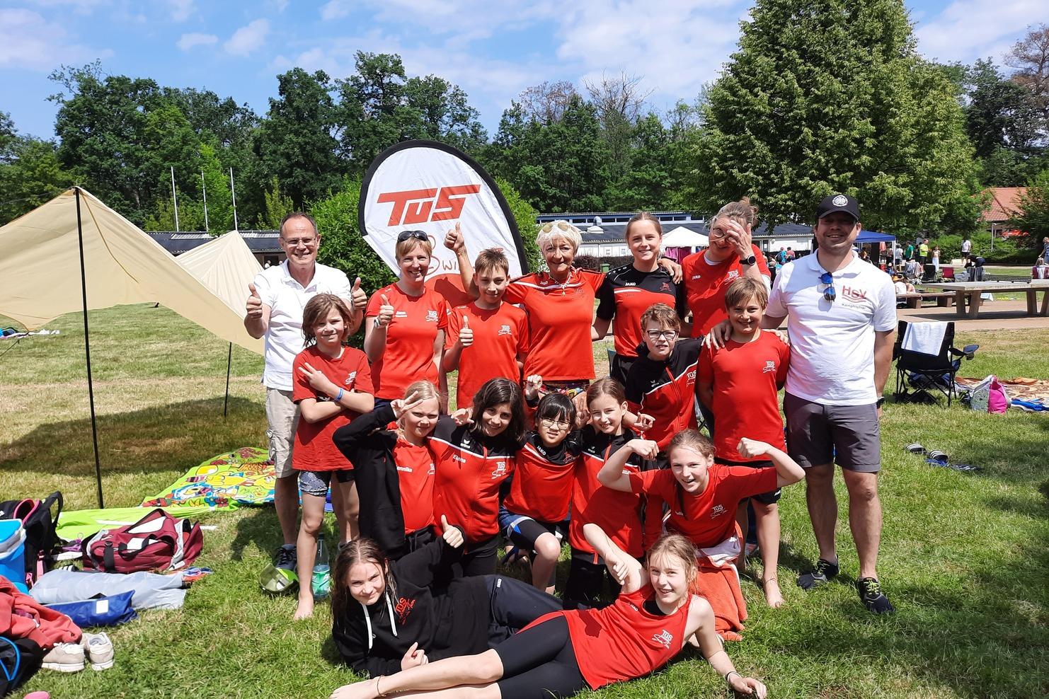 Gruppe von Kindern und Betreuern in roten T-Shirts, lächelnd im Freien vor Zelt und Bäumen, einige zeigen Daumen hoch.