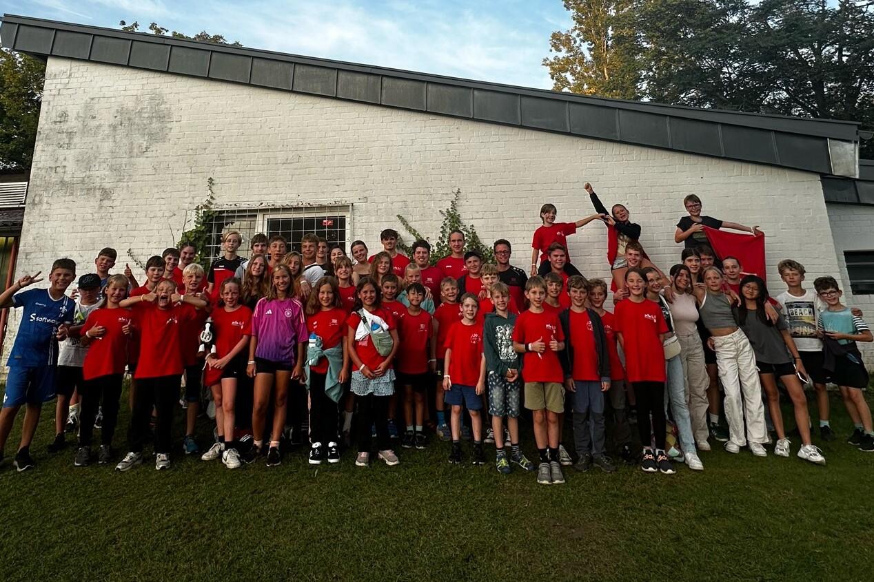 Gruppenfoto von etwa 60 Kindern und Jugendlichen in roten T-Shirts vor einem weißen Gebäude, einige machen lustige Posen.
