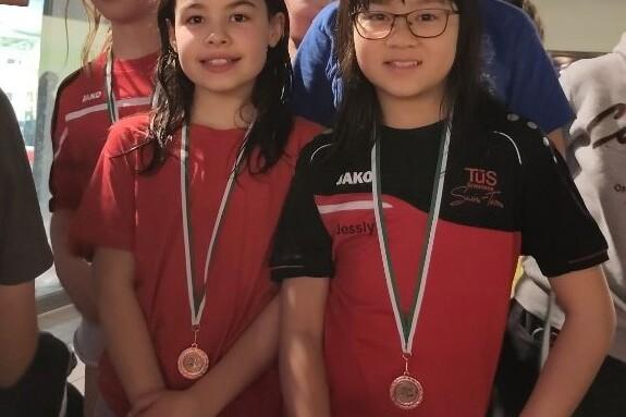 Vier Kinder in Sportkleidung mit Medaillen stehen lächelnd an einem Schwimmbad. Zwei Mädchen sind im Vordergrund.