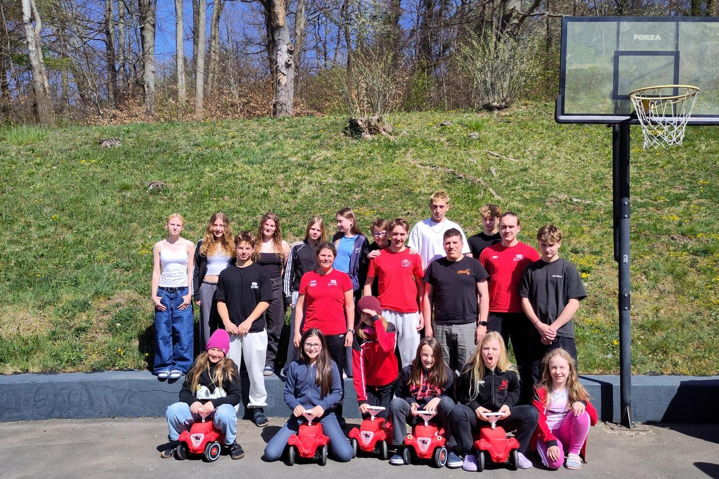 Gruppe von Jugendlichen posiert im Freien mit roten Spielautos vor einem Basketballkorb und einer grünen Wiese.