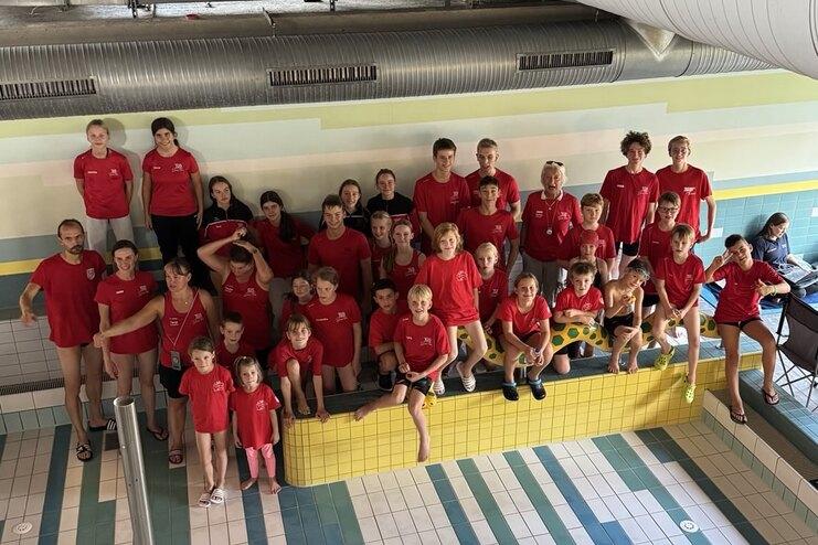 Gruppe von Schwimmern in roten Shirts posiert auf einer gelben Bank am Schwimmbecken in einem Freizeitbad.