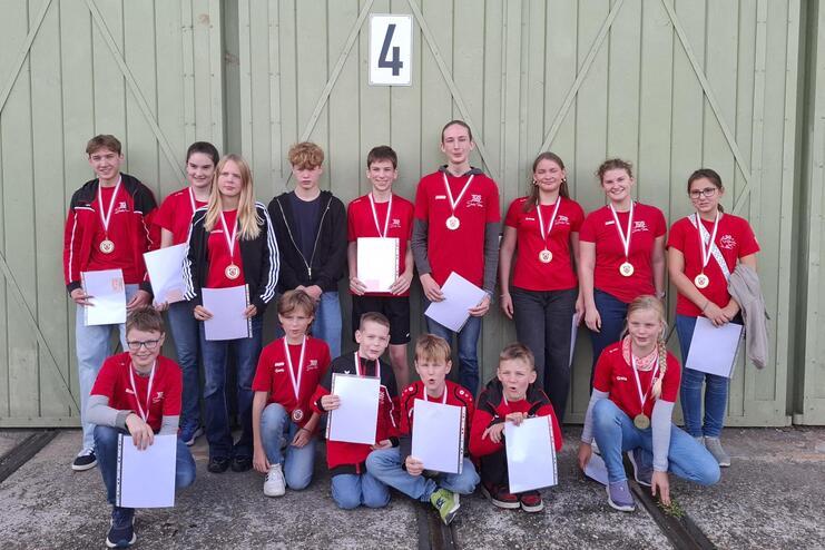 Gruppe von Jugendlichen in roten T-Shirts mit Medaillen, posierend vor einer grünen Wand, halten Urkunden in der Hand.