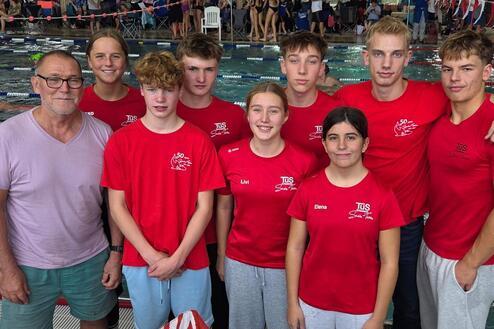 Gruppe von sieben Schwimmern in roten T-Shirts, stehend vor einem Schwimmbecken in einer Sporthalle.