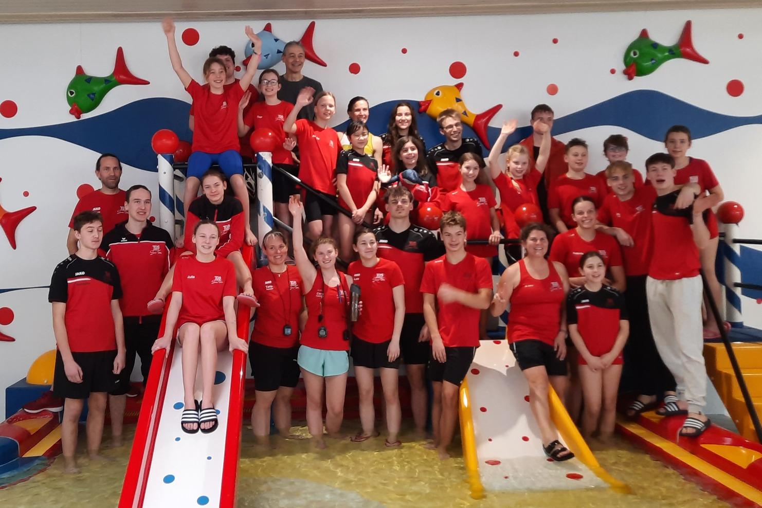 Gruppensfoto von Kindern und Betreuern in roten T-Shirts neben Schwimmrutschen in einem Pool, umgeben von bunten Wandmotiven.