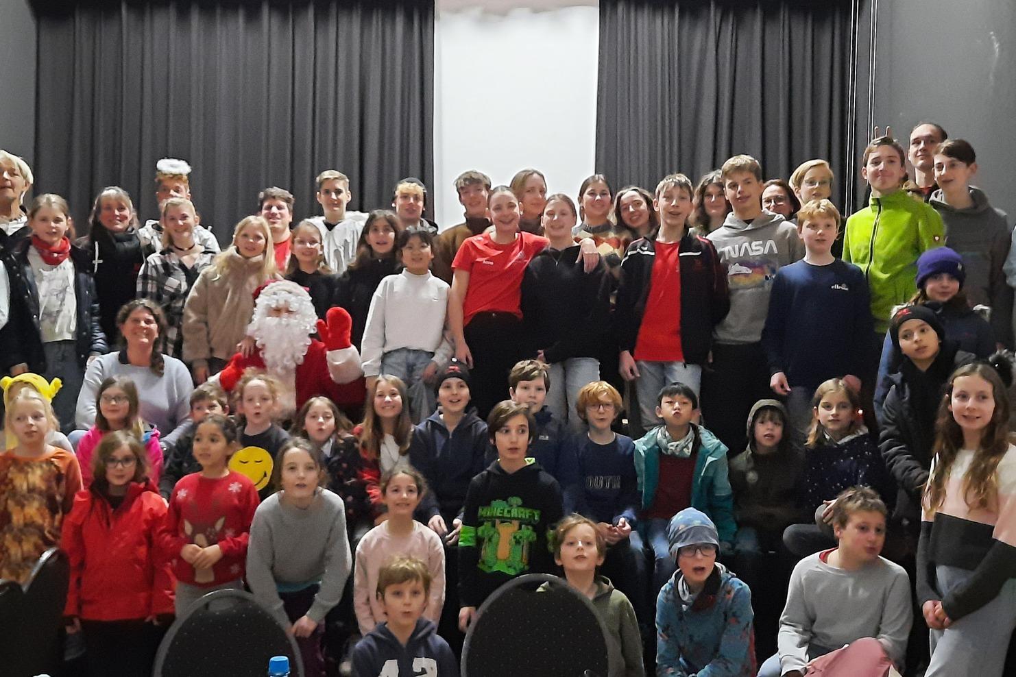 Gruppe von Kindern und Erwachsenen posiert festlich vor einem Weihnachtsbaum und einer Bühne mit Weihnachtsmann.