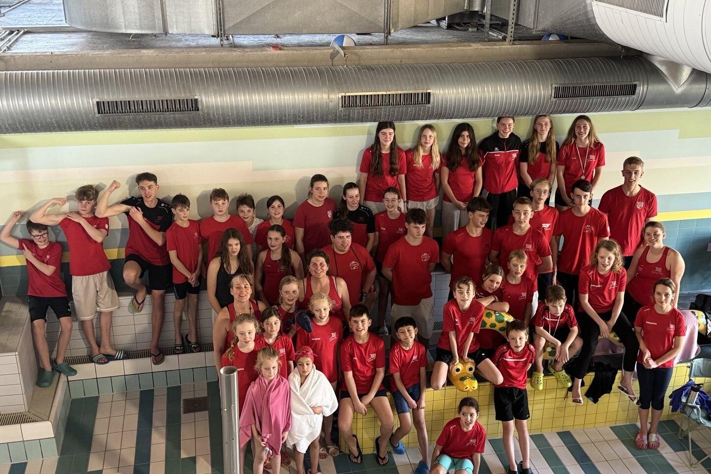 Gruppenshot von 44 Schwimmern in roten Shirts, lächelnd vor einer bunten Wand im Schwimmbad.