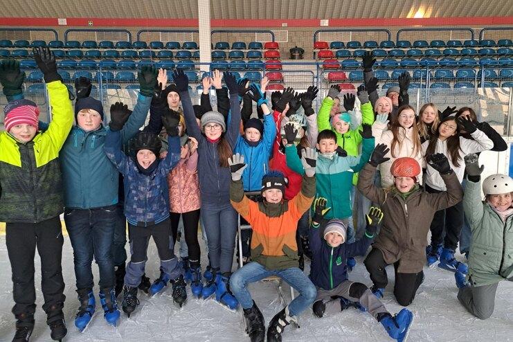 Gruppe von Kindern in Schlittschuhen hebt freudig die Hände auf einer Eisfläche in einer Eishalle.