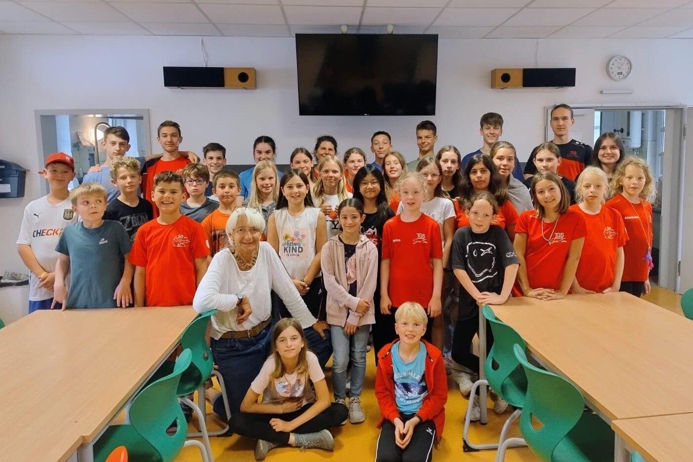 Gruppensfoto von 32 Personen in einem Klassenraum, einige tragen rote T-Shirts, eine ältere Frau sitzt vorn.