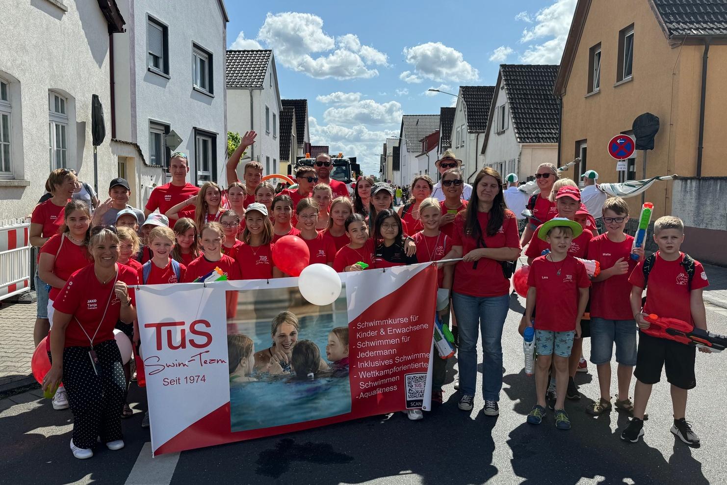Gruppe von Kindern und Erwachsenen in roten T-Shirts mit Banner bei einem Umzug auf einer sonnigen Straße.
