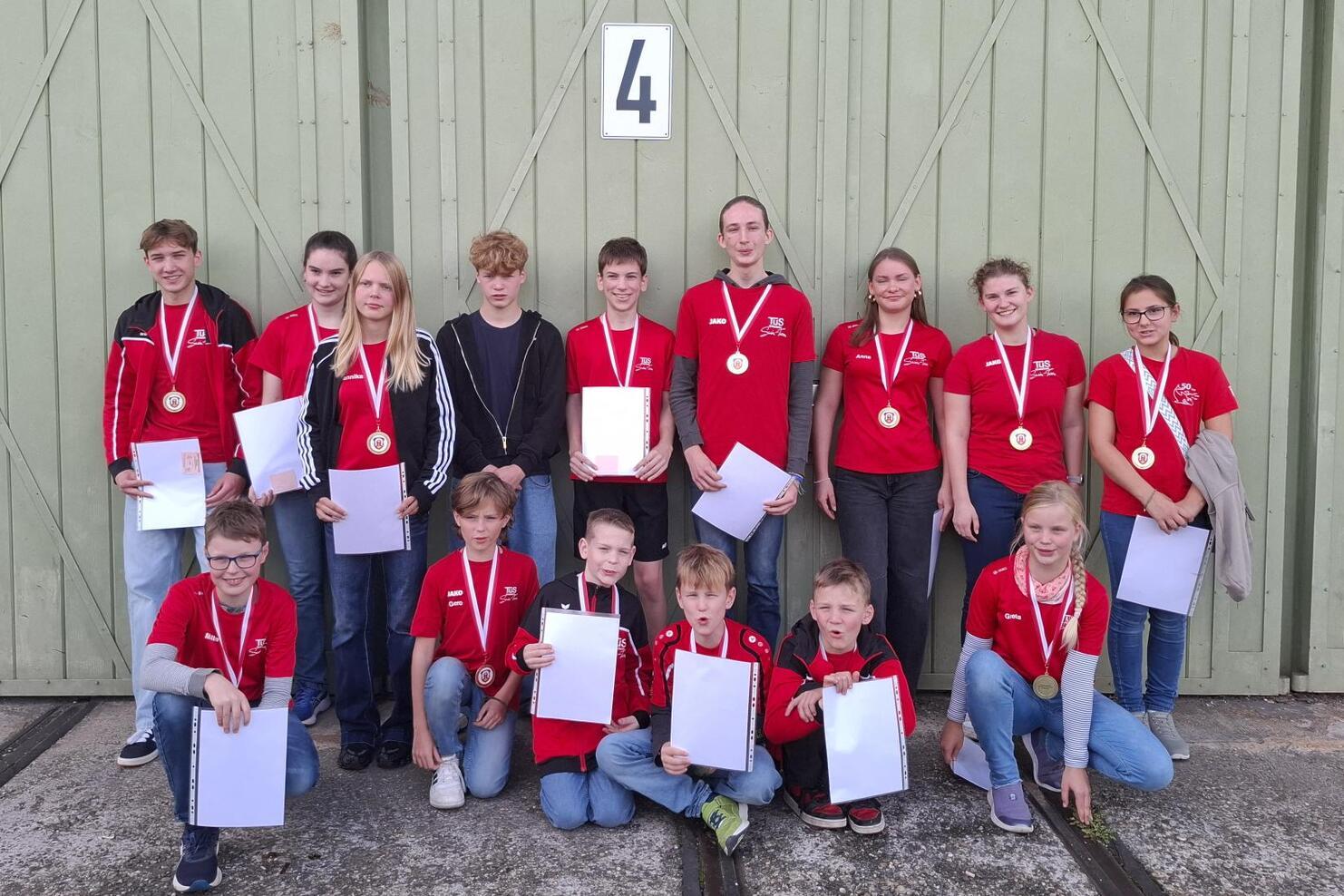 Gruppe von Jugendlichen in roten T-Shirts mit Medaillen, posierend vor einer grünen Wand, halten Urkunden in der Hand.
