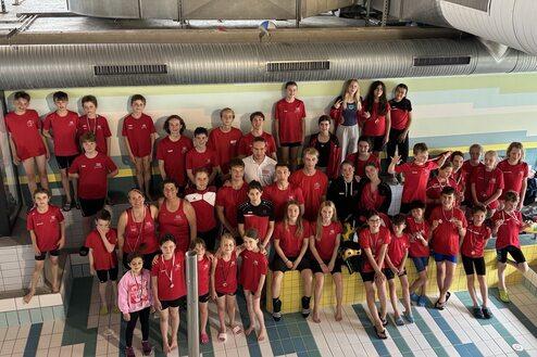 Gruppe von Schwimmern in roten T-Shirts posiert lächelnd am Beckenrand in einem Hallenbad.