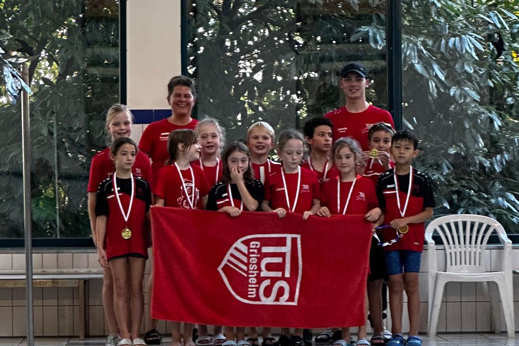 Gruppe junger Schwimmer in roten T-Shirts mit Medaillen, posiert vor einer Schwimmhalle mit einer Fahne.