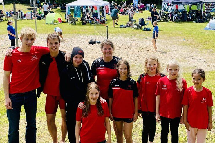 Gruppe junger Sportler in roten Trikots posiert auf einer Wiese mit Zelten und Zuschauern im Hintergrund unter blauem Himmel.