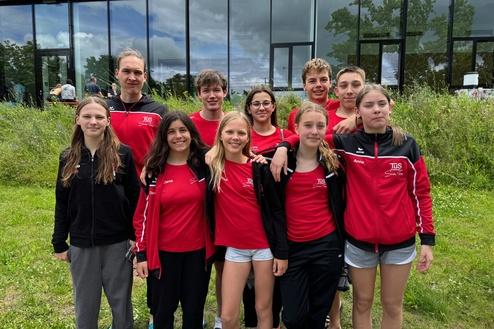 Gruppe von zehn Jugendlichen in roten Sportanzügen, stehend auf einer Wiese vor einem modernen Gebäude.