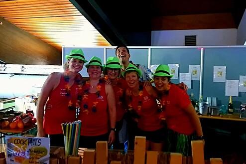 Gruppenshot von sechs Personen in bunten Shirts und grünen Hüten, umgeben von Cocktail-Zutaten an einer Bar.