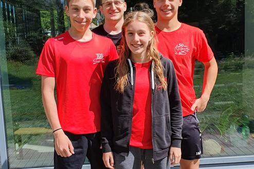 Vier Jugendliche in roten T-Shirts stehen vor einem großen Fenster und lächeln in die Kamera. Sonnenlicht und grüne Bäume im Hintergrund.
