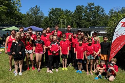 Gruppe von Schwimmern in roten T-Shirts steht auf einer Wiese, im Hintergrund Bäume und Zelte unter blauem Himmel.