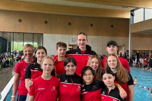 Gruppe junger Schwimmer in roten Trainingsanzügen, lächelnd vor einem Schwimmbecken und Holzstruktur im Hintergrund.