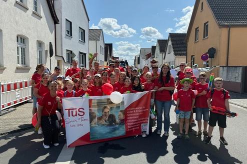 Gruppe von Kindern und Erwachsenen in roten T-Shirts mit Banner bei einem Umzug auf einer sonnigen Straße.