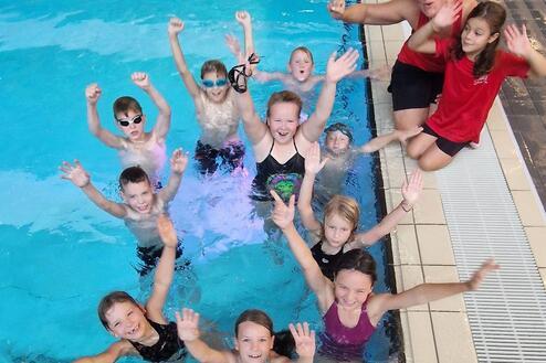 Gruppe fröhlicher Kinder und Trainerin posiert im Schwimmbecken, einige heben die Hände in die Luft.