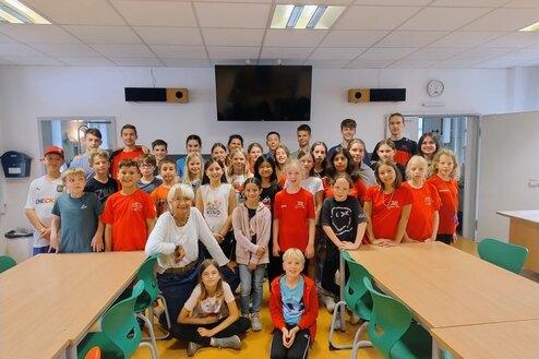 Gruppensfoto von 32 Personen in einem Klassenraum, einige tragen rote T-Shirts, eine ältere Frau sitzt vorn.