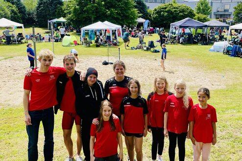 Gruppe junger Sportler in roten Trikots posiert auf einer Wiese mit Zelten und Zuschauern im Hintergrund unter blauem Himmel.