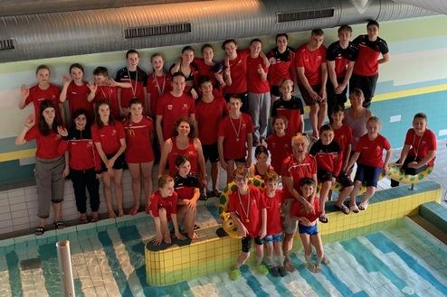 Gruppensfoto von Schwimmern in roten Shirts, stehend und sitzend am Beckenrand eines Schwimmbades.