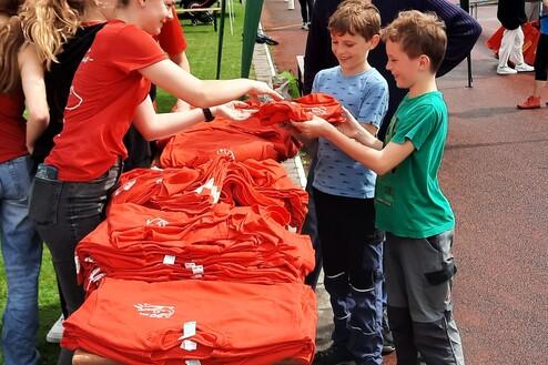 Zwei Kinder erhalten rote T-Shirts von einer jungen Frau an einem Tisch auf einem Sportgelände.
