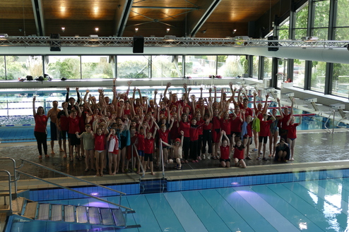 Gruppenfoto von Schwimmern in roten T-Shirts, die jubelnd am Beckenrand eines Schwimmbads stehen.