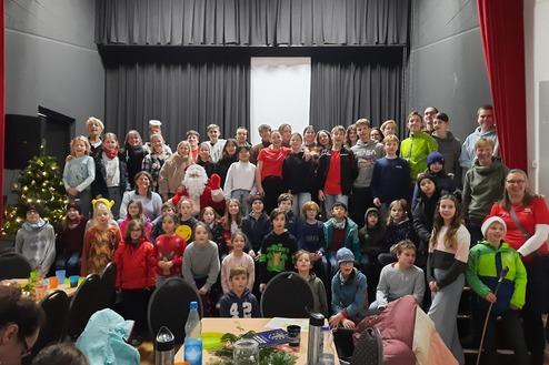 Gruppe von Kindern und Erwachsenen posiert festlich vor einem Weihnachtsbaum und einer Bühne mit Weihnachtsmann.