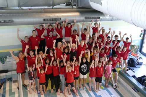 Gruppennfoto von Schwimmern in roten T-Shirts, die mit erhobenen Händen vor einem Schwimmbad posieren.