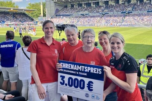 Vier Personen halten ein Schild mit der Aufschrift &bdquo;Ehrenamtspreis 3.000 &euro;&ldquo; in einem Fu&szlig;ballstadion. Hintergrund: Spielszene.