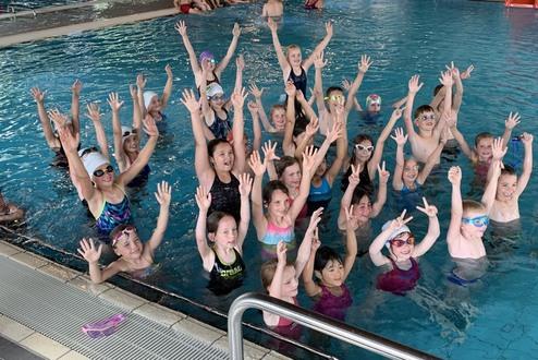 Gruppenaufnahme von Kindern im Schwimmbecken, die fröhlich die Arme heben und schwimmen.