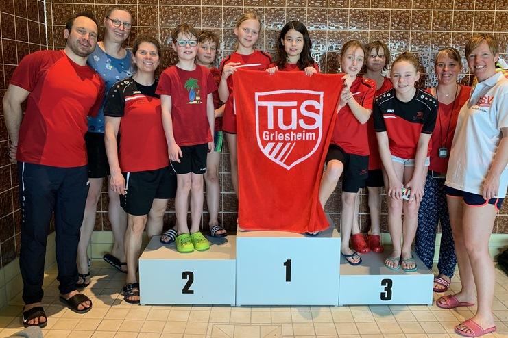 Gruppe von Schwimmern in roten Shirts posiert mit einer Medaille und einem Handtuch auf einem Siegerpodest.