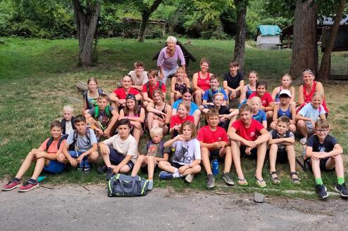 Gruppe von etwa 30 Kindern und Betreuern sitzend im Freien, umgeben von B&auml;umen und Wiese. Angenehme Sommersituation.