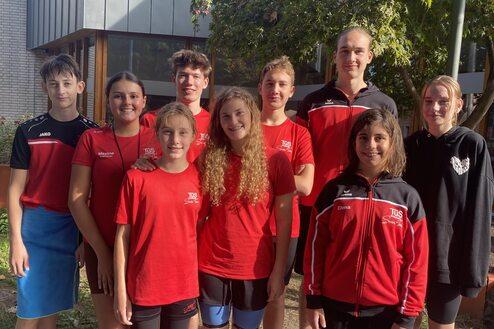 Gruppe von neun Jugendlichen in roten T-Shirts, die vor einem Geb&auml;ude stehen, umgeben von B&auml;umen und Blumen.