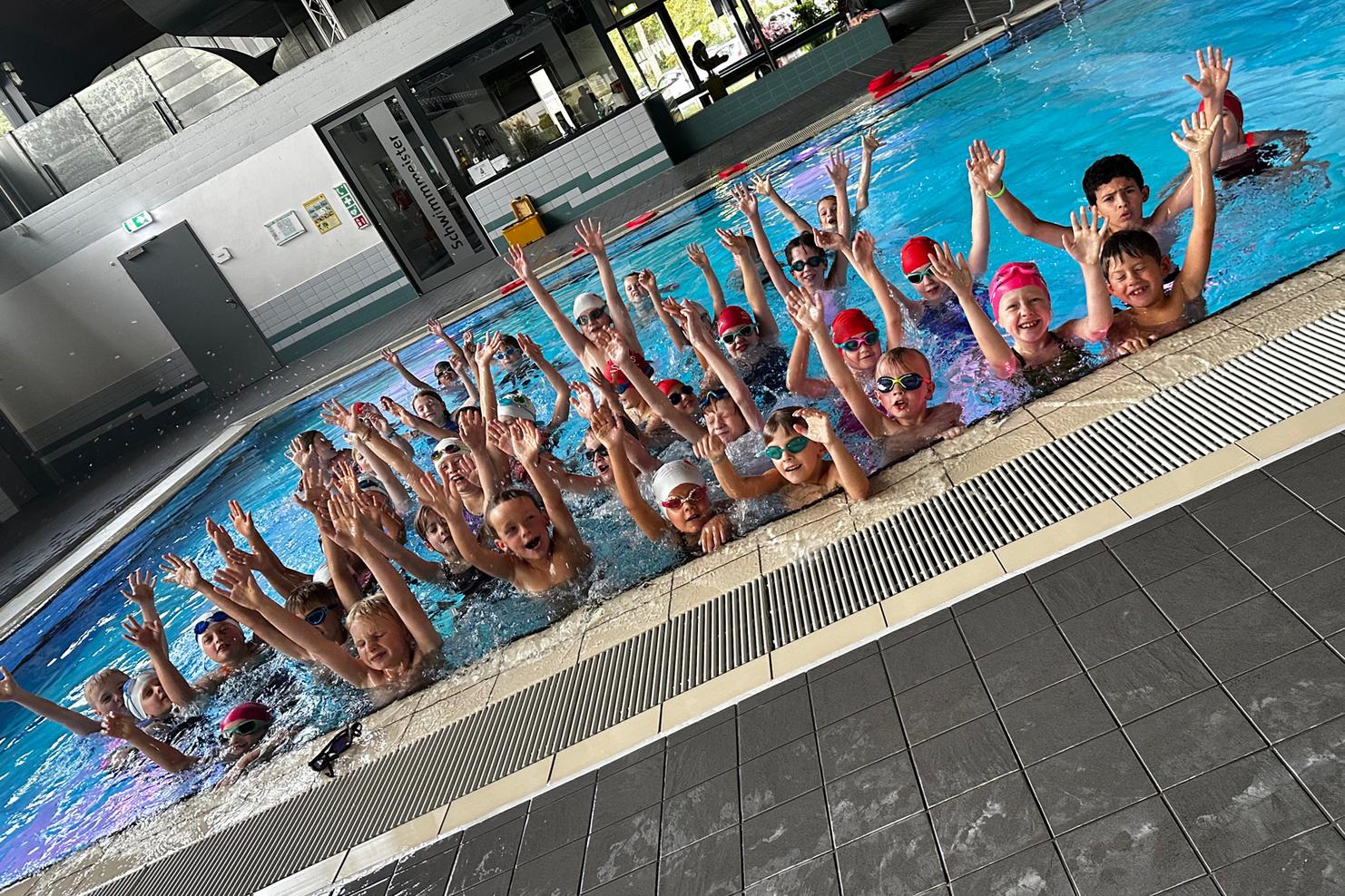 Mehrere Kinder mit Schwimmbrillen und Badekappen stehen im Wasser und heben die Hände in einem Schwimmbad.