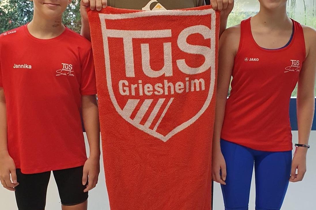 Drei Kinder in Sportkleidung stehen vor einem Fenster, einer hält ein Handtuch mit dem Logo von TuS Griesheim.