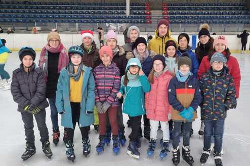 Gruppe von Kindern und Erwachsenen in Winterkleidung auf einer Eislauffl&auml;che, alle tragen Schlittschuhe.