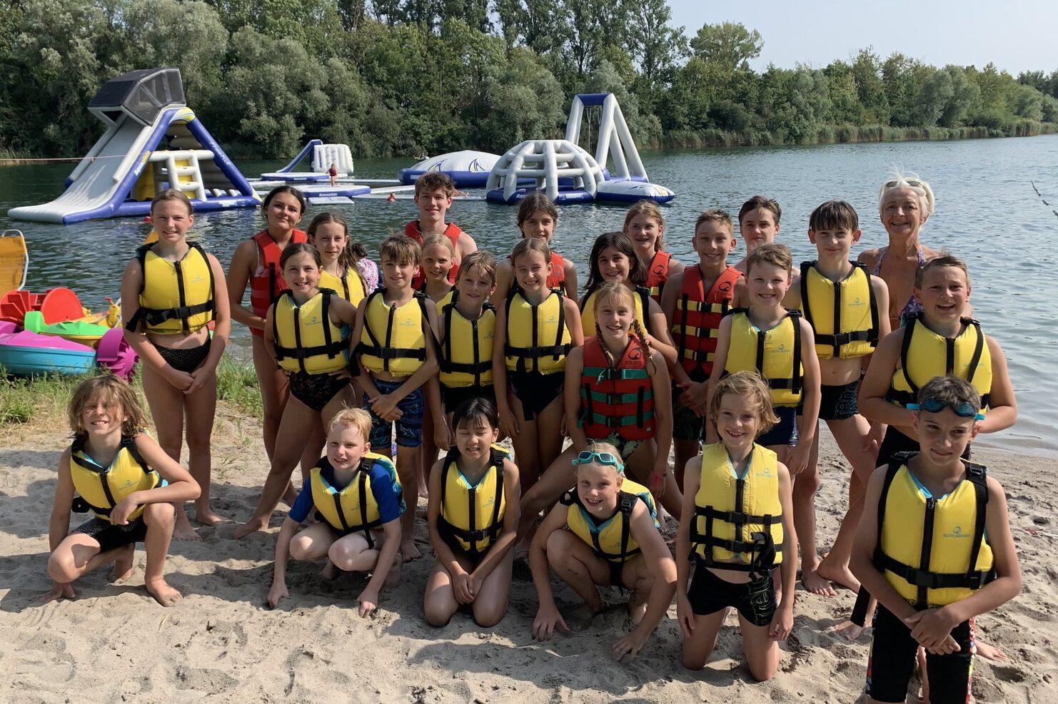 Gruppe von Kindern in Schwimmwesten am Strand, mit Wasserattraktionen im Hintergrund. Sonniger Tag am See.