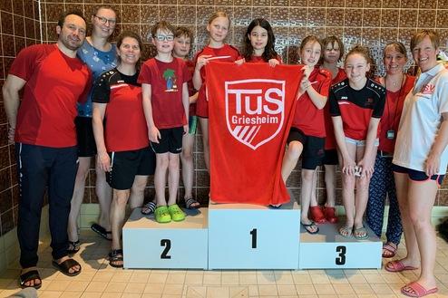 Gruppe von Schwimmern in roten Shirts posiert mit einer Medaille und einem Handtuch auf einem Siegerpodest.