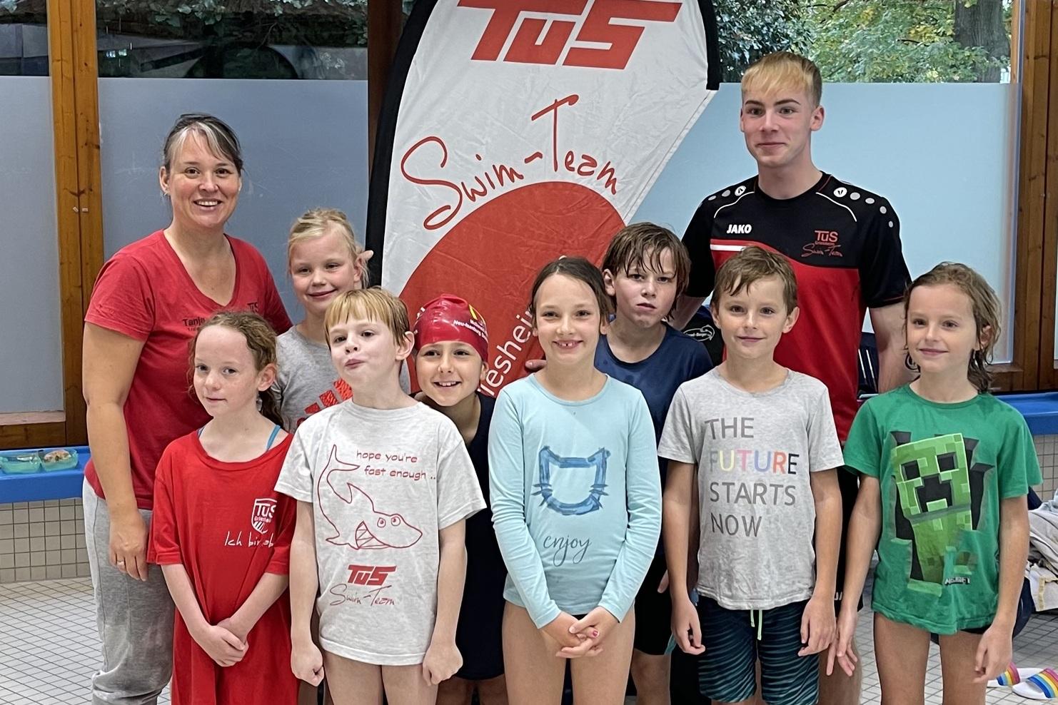 Gruppe von Kindern und Trainern vor einem Swim-Team-Schild in einem Hallenbad, umgeben von Wasser und Schwimmausrüstung.