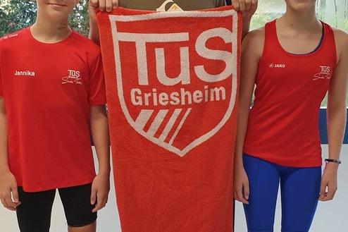 Drei Kinder in Sportkleidung stehen vor einem Fenster, einer h&auml;lt ein Handtuch mit dem Logo von TuS Griesheim.
