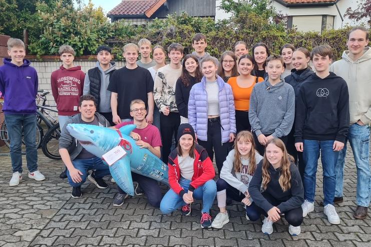 Gruppe von Jugendlichen posiert mit einem aufblasbaren Hai vor einer Backsteinmauer und Fahrrädern im Hintergrund.