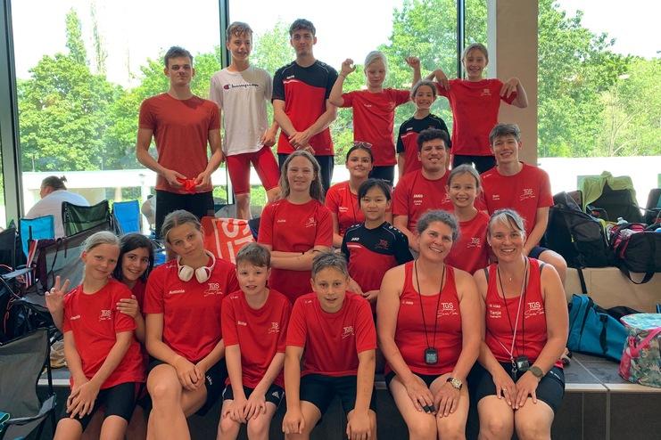 Gruppe von Schwimmern und Trainern in roten T-Shirts, lächelnd vor großen Fenstern mit Blick auf die Natur.
