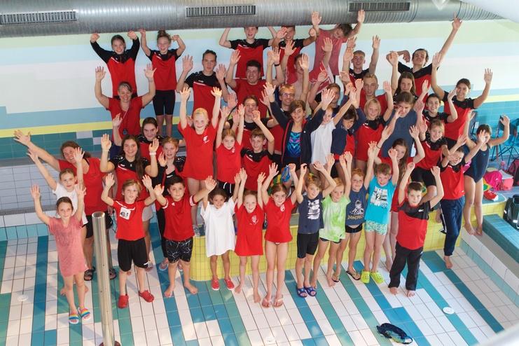 Gruppenshot von Schwimmern in roten Shirts, die fröhlich mit erhobenen Händen in einem Hallenbad stehen.
