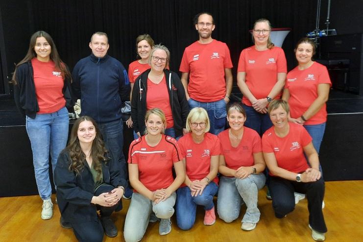 Gruppe von 12 Personen in roten T-Shirts, die lächeln und auf einer Bühne in einem hellen Raum stehen.