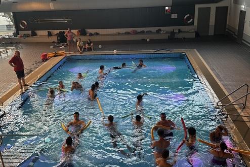 Gruppe von Schwimmern mit Noodles in einem Innenpool, umgeben von großen Fenstern und einer Uhr an der Wand.
