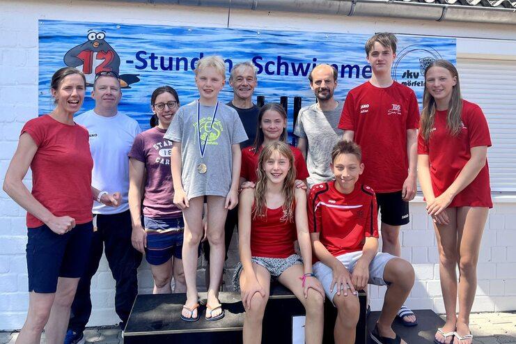 Gruppennaufnahme von Schwimmern auf dem Treppchen nach einem Wettkampf mit Medaillen und einer Banner im Hintergrund.