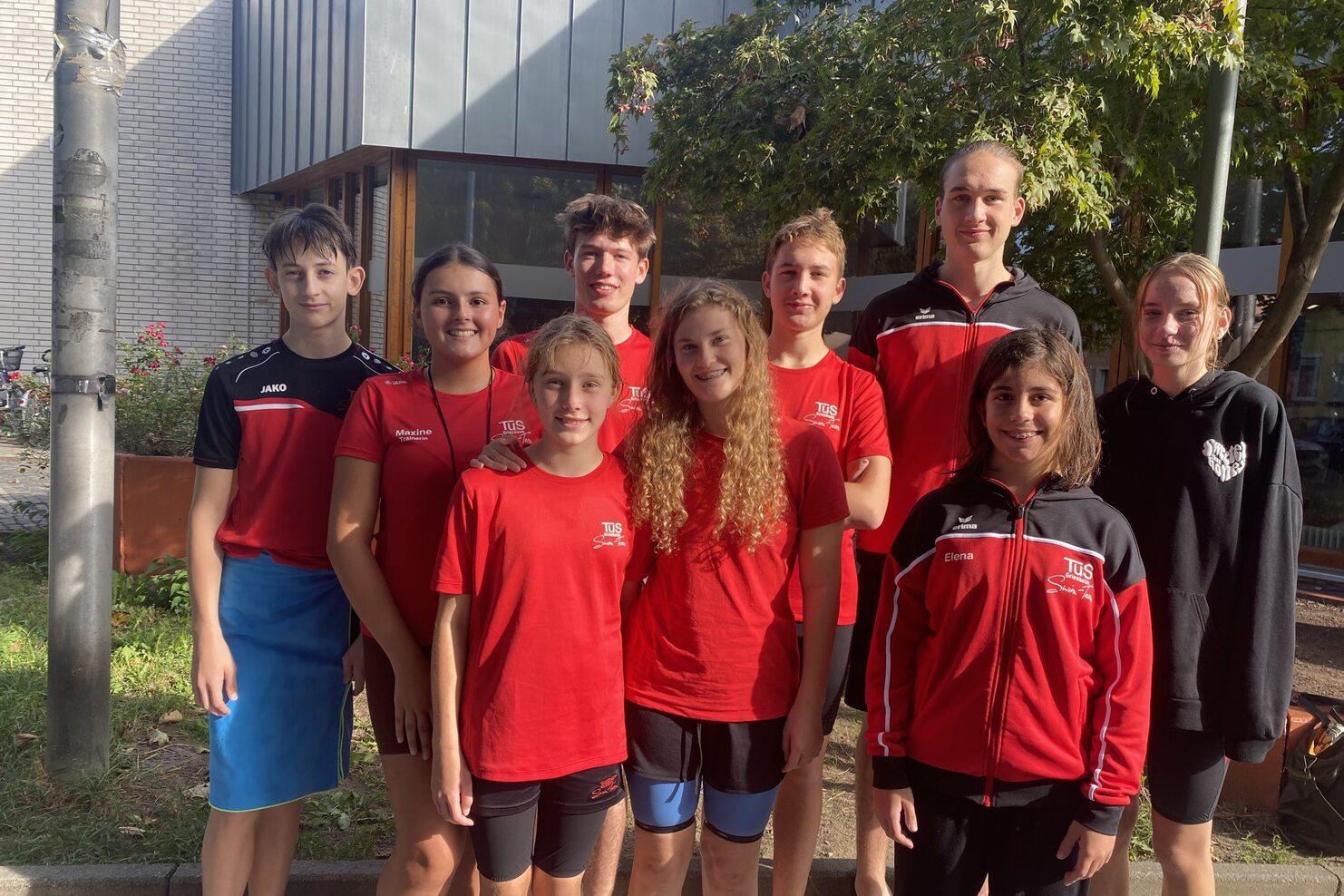 Gruppe von neun Jugendlichen in roten T-Shirts, die vor einem Gebäude stehen, umgeben von Bäumen und Blumen.
